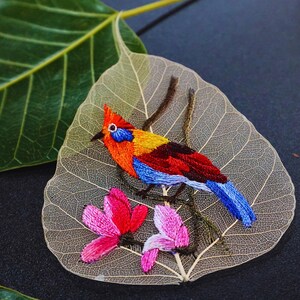 Puede incluir: Un pájaro bordado de colores vivos posado en una rama con flores rosas, todo cosido sobre una hoja delicada y translúcida.