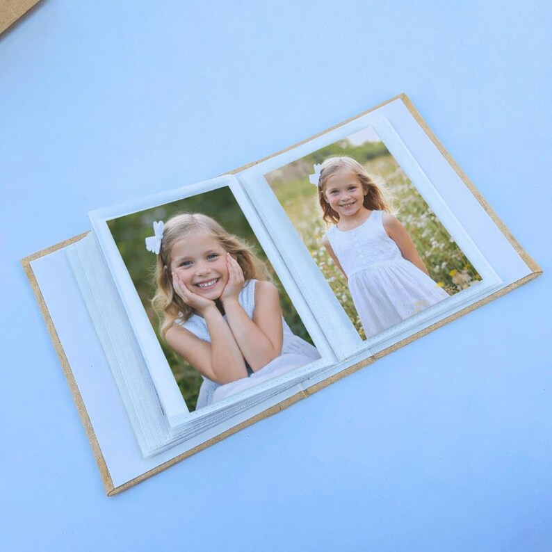 May include: An open photo album with two framed photos of a young girl in a white dress. The album has a light brown cover and white pages. The girl is smiling in both photos, one with her hands on her face.