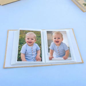 May include: Open photo album with two baby photos. Each photo features a smiling baby wearing a light blue striped onesie. The album has a light brown cover and white pages. The background is a light blue surface.