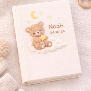 May include: A cream-colored baby memory book with a teddy bear, moon, and star design. The book cover features the name "Noah" and the date "04.16.24" in a soft, embroidered style. The design includes a teddy bear holding a star, sitting on a cloud.