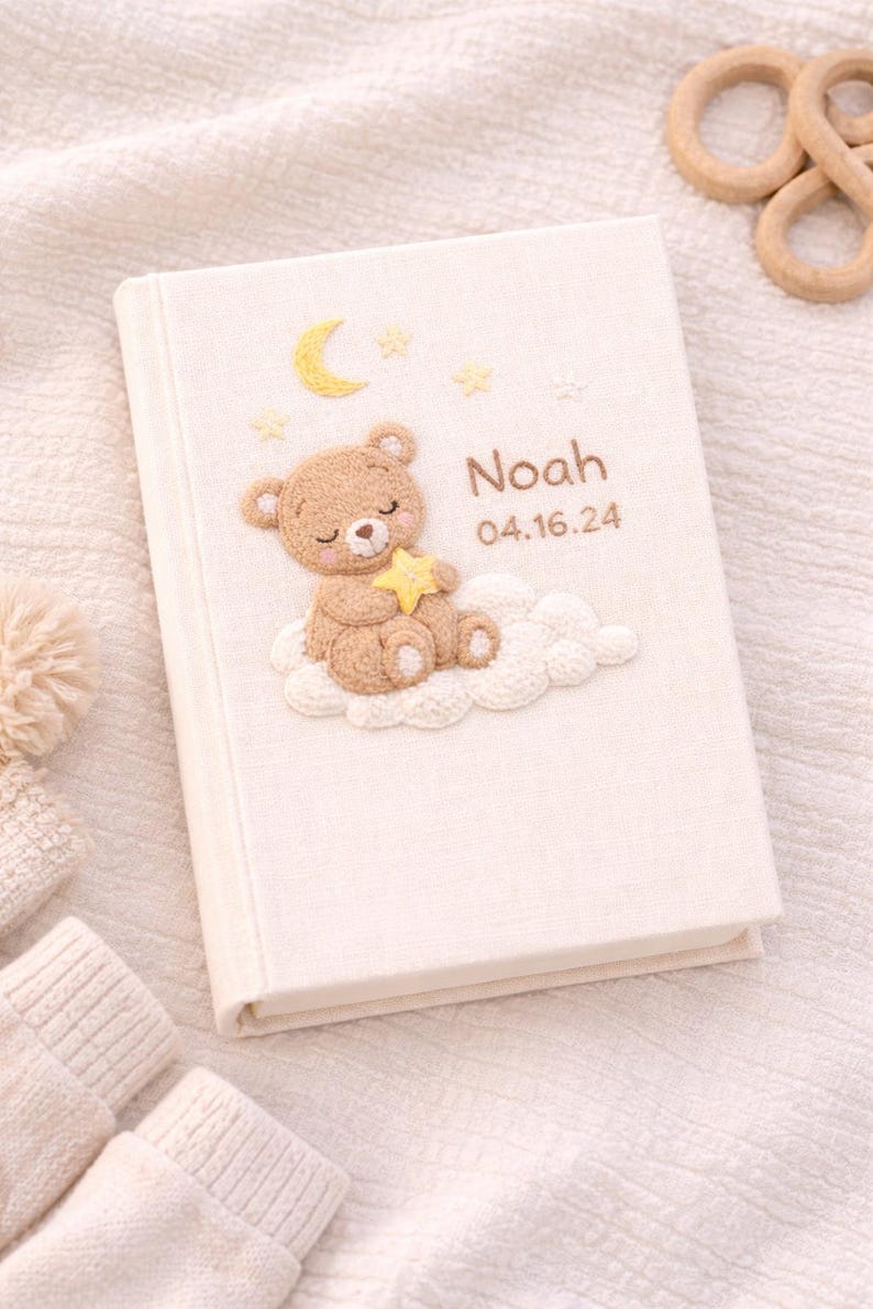 May include: A cream-colored baby memory book with a teddy bear, moon, and star design. The book cover features the name "Noah" and the date "04.16.24" in a soft, embroidered style. The design includes a teddy bear holding a star, sitting on a cloud.