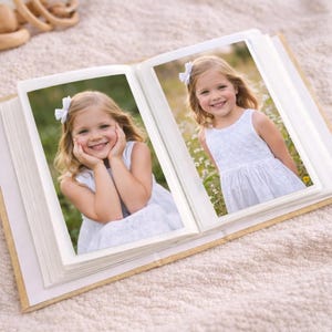 May include: An open photo album with two pictures of a young girl in a white dress and a white bow in her hair. The album is open on a beige blanket. The girl is smiling in both photos.