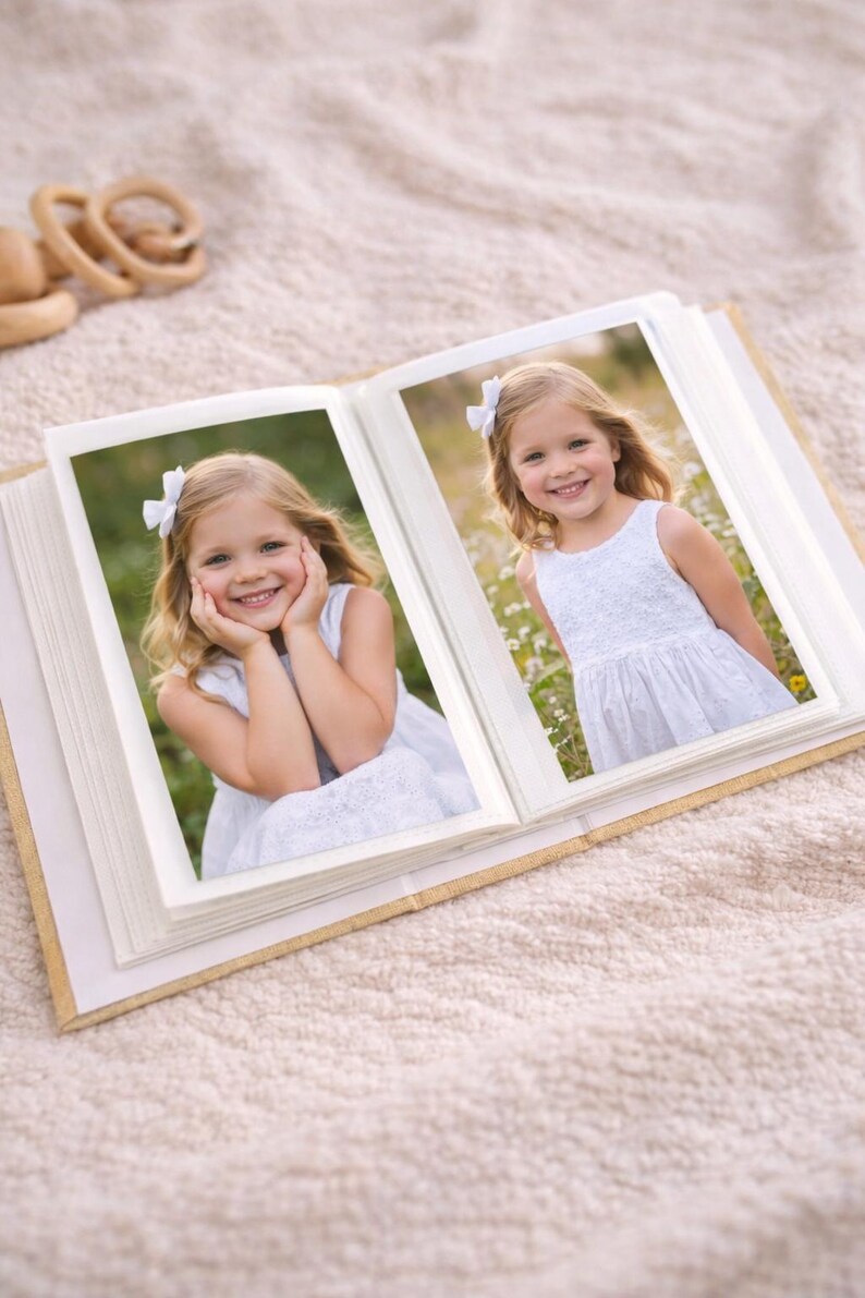 May include: An open photo album with two pictures of a young girl in a white dress and a white bow in her hair. The album is open on a beige blanket. The girl is smiling in both photos.
