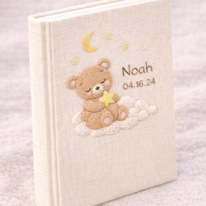 May include: A cream-colored baby memory book with an embroidered design. The cover features a sleeping teddy bear on a cloud, a crescent moon, stars, and the name "Noah" with the date 04.16.24.