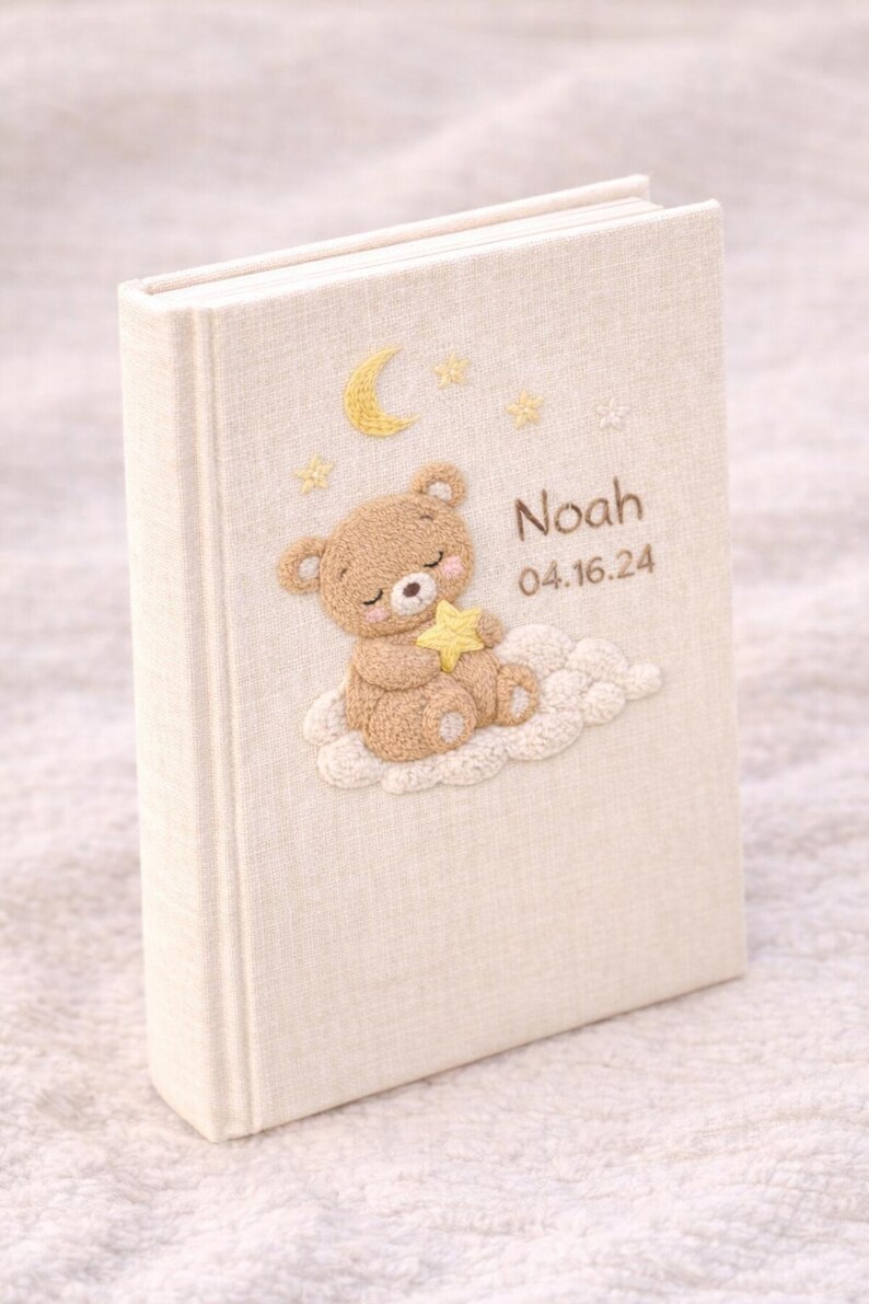 May include: A cream-colored baby memory book with an embroidered design. The cover features a sleeping teddy bear on a cloud, a crescent moon, stars, and the name "Noah" with the date 04.16.24.