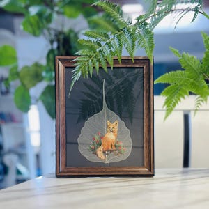 May include: A framed artwork featuring a fox sitting on a delicate, translucent leaf. The leaf is surrounded by small red flowers and the fox is depicted in shades of orange and white.