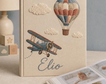 May include: A beige fabric-covered photo album featuring embroidered designs of a blue biplane, a multi-colored hot air balloon, and white clouds. The name Elio is embroidered in blue script at the bottom. The album is open, displaying photos.