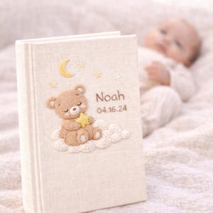 May include: A beige baby memory book with an embroidered teddy bear holding a star, sitting on a cloud. The book is personalized with the name "Noah" and the date "04.16.24". The cover also features embroidered stars and a crescent moon.