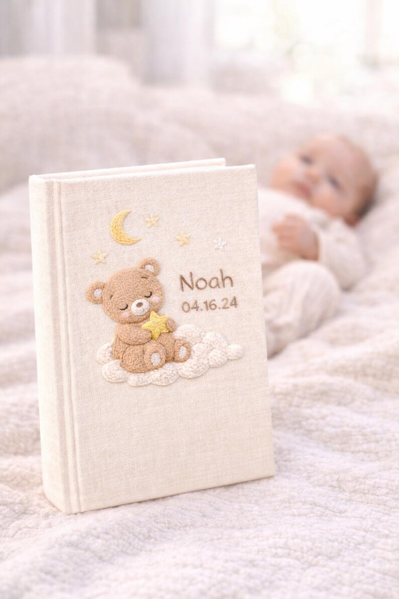 May include: A beige baby memory book with an embroidered teddy bear holding a star, sitting on a cloud. The book is personalized with the name "Noah" and the date "04.16.24". The cover also features embroidered stars and a crescent moon.