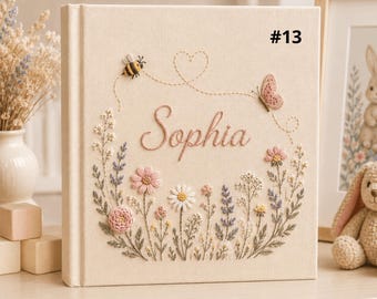 May include: A cream-colored fabric-covered book featuring embroidered floral designs, a bee, and a butterfly. The name "Sophia" is stitched in the center in a cursive font, with the text "#13" visible in the upper right corner of the cover.