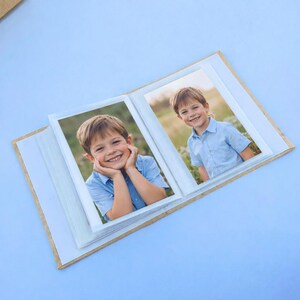 May include: A small photo album with two pictures of a smiling child in a blue shirt. The album has a cardboard cover and white photo frames. The background is a light blue surface.