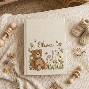 May include: A cream-colored baby memory book with the name "Oliver" embroidered on the cover. The cover features a brown teddy bear, flowers, butterflies, and bees. A pair of knitted baby booties and a wooden rattle are also visible.