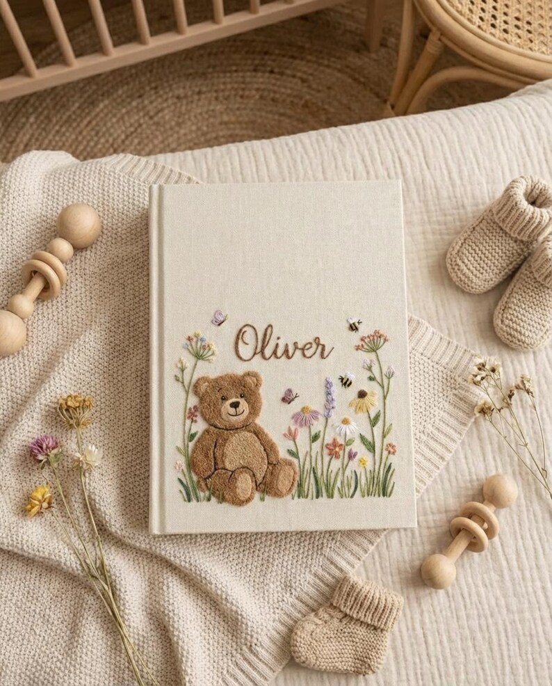 May include: A cream-colored baby memory book with the name "Oliver" embroidered on the cover. The cover features a brown teddy bear, flowers, butterflies, and bees. A pair of knitted baby booties and a wooden rattle are also visible.