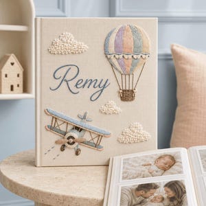 Könnte beinhalten: Ein beigefarbenes, stoffbezogenes Fotoalbum mit gestickten Motiven eines pastellfarbenen Heißluftballons, eines blauen Doppeldeckers und strukturierten weißen Wolken. Der Name Remy ist in blauer Schreibschrift auf dem Einband gestickt.