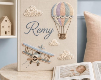 May include: A beige fabric-covered photo album featuring embroidered designs of a pastel hot air balloon, a blue biplane, and textured white clouds. The name Remy is embroidered in blue script on the front cover, which stands upright.