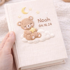 May include: A beige fabric-covered baby memory book with an embroidered design. The design features a sleeping teddy bear on a cloud, a crescent moon, stars, and the name "Noah" with the date 04.16.24.