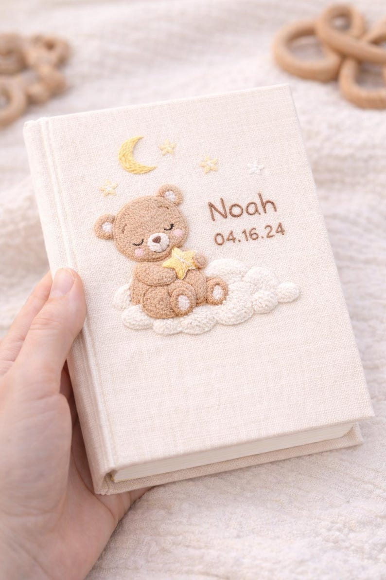 May include: A beige fabric-covered baby memory book with an embroidered design. The design features a sleeping teddy bear on a cloud, a crescent moon, stars, and the name "Noah" with the date 04.16.24.