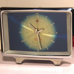 May include: A rectangular, chrome-framed alarm clock with a blue and yellow gradient face. The clock features silver hands and numerals, with the text "SHANGHAI" visible on the face. The clock has a retro design.