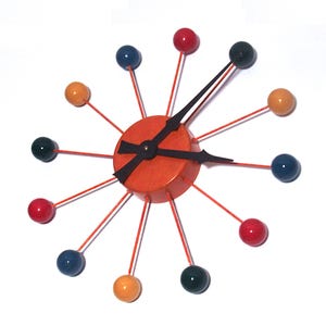 Puede incluir: Un reloj de pared de estilo retro con una esfera naranja vibrante y manecillas negras. El reloj presenta esferas coloridas en rojo, azul, verde y amarillo, conectadas por finas varillas naranjas. El diseño es de estética moderna de mediados de siglo.