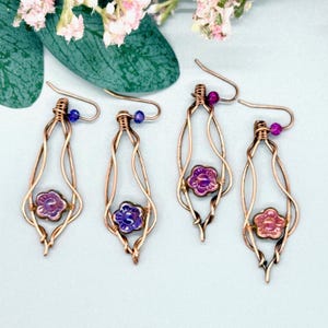 Könnte beinhalten: Vier Paar handgefertigte Ohrringe aus Kupferdraht. Jeder Ohrring hat eine Tropfenform mit einer blütenförmigen Perle in der Mitte. Die Blumen sind lila und rosa, mit kleinen blauen und lila Perlen oben.