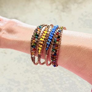 Puede incluir: Cuatro pulseras de alambre de cobre con cuentas coloridas se muestran en una muñeca. Las pulseras presentan cuentas amarillas, azules y multicolores. El alambre está envuelto alrededor de las cuentas, creando un efecto decorativo.