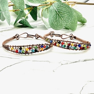 May include: Two copper wire bracelets with colorful beads. The bracelets have a hook and eye closure. The beads are in a rainbow of colors, including red, orange, yellow, green, blue, and purple. The bracelets are displayed on a white marble surface.