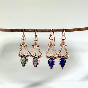 May include: Four pairs of handcrafted copper wire earrings. Two pairs feature teardrop-shaped green and purple gemstones, while the other two showcase blue gemstones. The earrings are suspended from a wooden rod against a white background.
