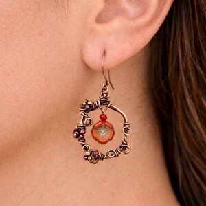 May include: A handcrafted copper wire earring with a circular design. The earring features a central orange flower-shaped bead, surrounded by smaller purple and gold beads. A small red bead hangs below the hook.