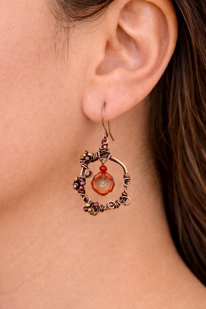 May include: A handcrafted copper wire earring with a circular design. The earring features a central orange flower-shaped bead, surrounded by smaller purple and gold beads. A small red bead hangs below the hook.