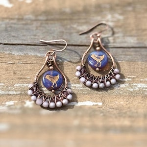 May include: A pair of teardrop-shaped earrings featuring copper wire frames, each centered with a round purple glass bead displaying a gold bird silhouette. The lower curve of each frame is wrapped with small, light-colored beads and dark wire accents.