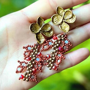 May include: A pair of gold-colored flower-shaped stud earrings with dangling beaded accents. The beaded section features red, white, and gold beads. The earrings are displayed on a hand against a blurred green background.