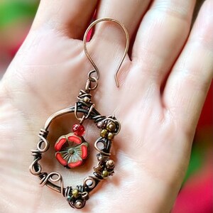 May include: A handcrafted copper earring with a floral design. The earring features a wire-wrapped frame with a red and green flower pendant and small, colorful beads. The hook is a simple, curved design.