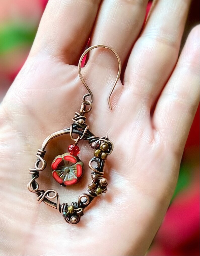 May include: A handcrafted copper earring with a floral design. The earring features a wire-wrapped frame with a red and green flower pendant and small, colorful beads. The hook is a simple, curved design.