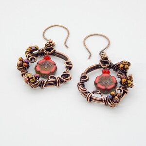 May include: Copper wire earrings with a floral design. Each earring features a red flower bead at the center, surrounded by copper wire and small, colorful beads. The earrings have a hook closure.
