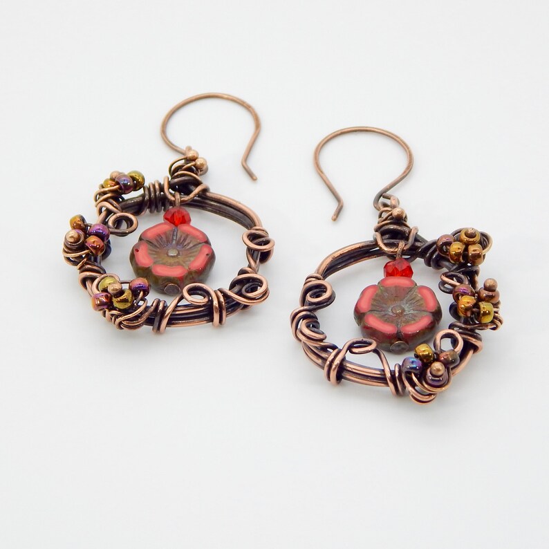 May include: Copper wire earrings with a floral design. Each earring features a red flower bead at the center, surrounded by copper wire and small, colorful beads. The earrings have a hook closure.