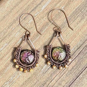 May include: A pair of copper wire-wrapped dangle earrings featuring a teardrop-shaped frame. Each earring contains a round, iridescent glass cabochon with swirling purple and green patterns, accented by small copper beads along the bottom edge.