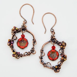 May include: Handmade copper wire earrings. Each earring features a square frame with intricate wire wrapping, small colorful beads, and a dangling red flower-shaped bead. The earrings have a hook closure and a rustic, artistic design.