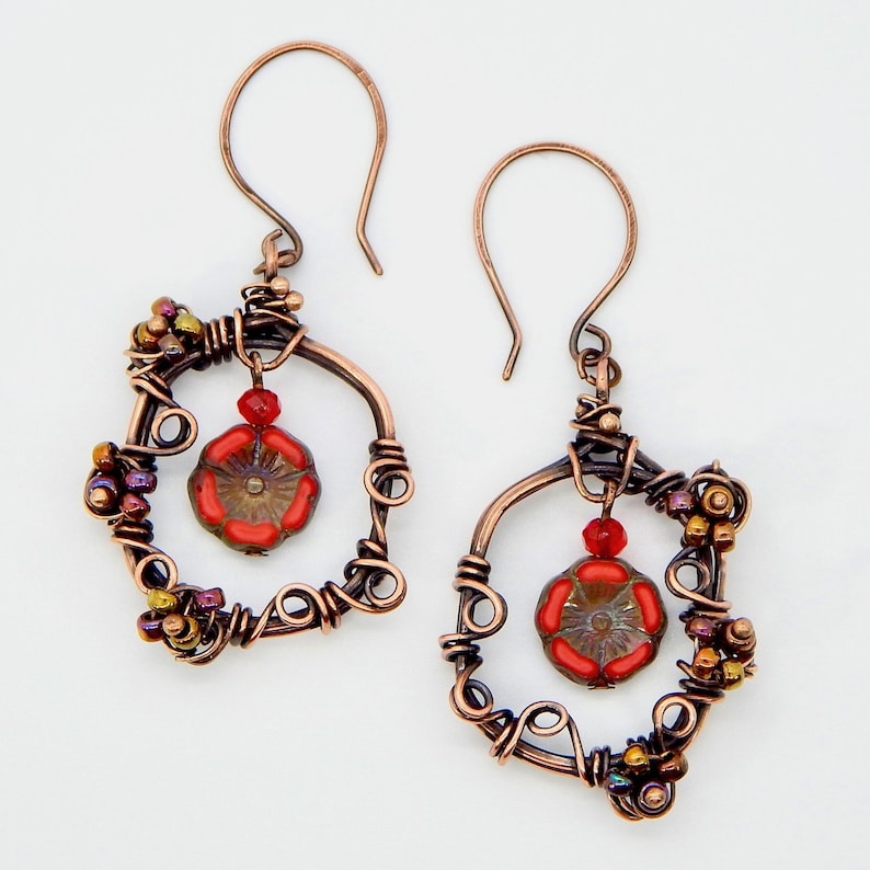 May include: Handmade copper wire earrings. Each earring features a square frame with intricate wire wrapping, small colorful beads, and a dangling red flower-shaped bead. The earrings have a hook closure and a rustic, artistic design.