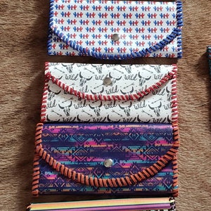 May include: Five different colorful wallets with various patterns. The wallets have a snap closure and are made of a leather-like material. The patterns include cacti, a skull with the word "wild", geometric shapes, and a leopard print.