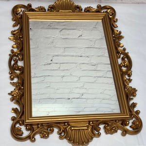 May include: A rectangular gold-colored mirror with an ornate, detailed frame. The frame features scrolling floral and shell-like designs. The mirror reflects a white brick wall. The overall style is vintage and decorative.