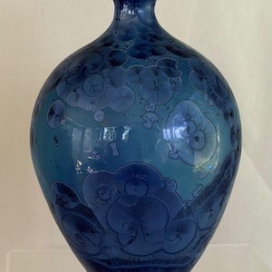 May include: A ceramic vase with a narrow neck and a rounded body, featuring a glossy finish in shades of deep blue and light blue. The surface is decorated with distinct crystalline patterns that resemble circular floral or star-like formations.