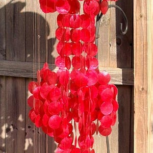 May include: A vibrant red wind chime with a decorative top and cascading circular elements. The outdoor decor hangs from a black metal frame, creating a visually appealing display. The chime is set against a wooden fence.