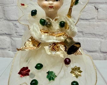 May include: A decorative angel figurine with a pale face and blonde hair, wearing a white sheer dress with gold trim. The dress is adorned with small red and green beads, metallic gold star-shaped accents, and a gold bell held in one hand.