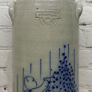 May include: A tall, cylindrical beige stoneware crock with two side handles. It features a blue hand-painted illustration of a bear decorating a Christmas tree. The crock is stamped with the text SALMON FALLS STONEWARE DOVER N.H. 1990.