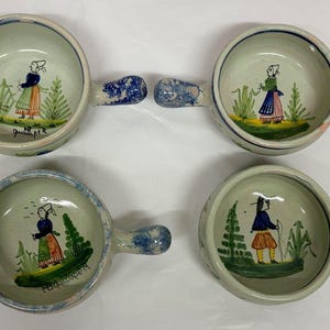 May include: Four hand-painted ceramic bowls with handles. Each bowl features a light green interior with a folk art illustration of a person in traditional clothing. The handles are blue and white. The bowls are approximately 15 cm in diameter.