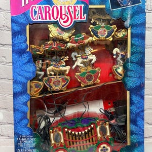 May include: A Holiday Carousel boxed set featuring a string of six carousel horses and a circus organ that plays 21 favorite Christmas carols. The blue packaging displays the product name, images of the carousel, and text describing the features.