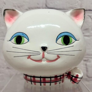 May include: A white ceramic cat head figurine featuring large green eyes with blue eyelids, a small black nose, and a pink mouth. The cat wears a red and black plaid collar around its neck, and the inner ears are painted with a soft pink hue.