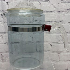 May include: A clear glass coffee pot with a glass lid and handle. The pot has a silver band around the middle and a red accent on the handle. The handle is made of clear glass. The pot is sitting on a clear surface.