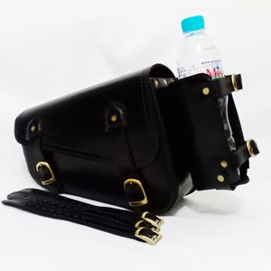 May include: Black leather motorcycle saddlebag with a gold buckle and a strap. The bag has a side pocket for a water bottle. The water bottle is clear plastic with a blue lid and a label that says "Miss B's And Drinks".