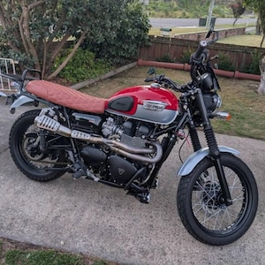 May include: A red and silver Triumph motorcycle with a brown leather seat and a black exhaust pipe. The motorcycle has a black front fender and a black rear fender. The motorcycle is parked on a gray driveway.